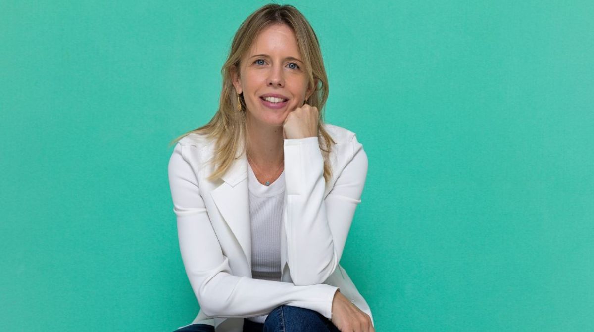 Cecília Antipoff posa para foto. Ela está vestida com um blazer branco, uma blusa branca e uma calça jeans. Ela é mulher branca, loira de olhos azuis. O fundo da foto é de cor verde água.