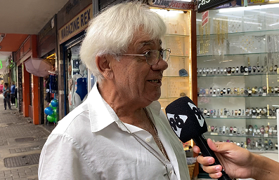 Um senhor de cabelos brancos fala ao microfone da 98, em frente a uma joalheria, sobre a Times Square mineira. No peito ele carrega um crucifixo dourado.