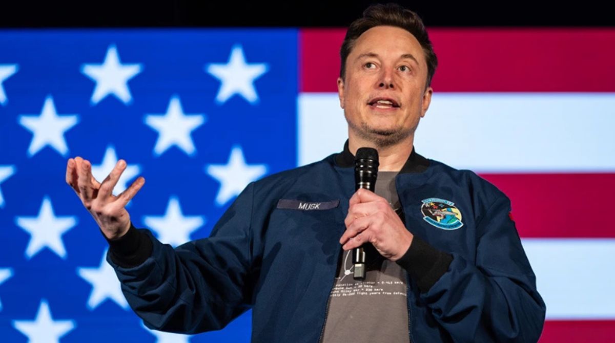 Elon Musk com microfone na mão e a bandeira dos Estados Unidos ao fundo