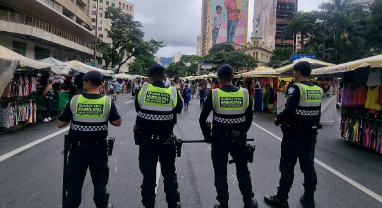 Quatro Guardas Municipais, fardados, são vistos de costas em uma avenida cheia de pessoas.